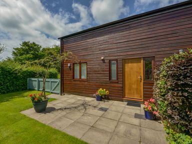 An exterior wooden wall with a wooden door and two windows a paved patio with potted flowers and a small tree a green lawn and a light green fence with trees in the background at Cholwell Barn Apartment Mary Tavy
