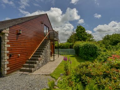 An exterior view of a building with wooden siding and stone walls with stairs and a garden with bushes and flowers at Cholwell Barn Apartment Mary Tavy