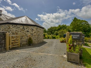 A stone building with a slate roof next to a gravel driveway and a sign that says Cholwell Barn with plants in Mary Tavy