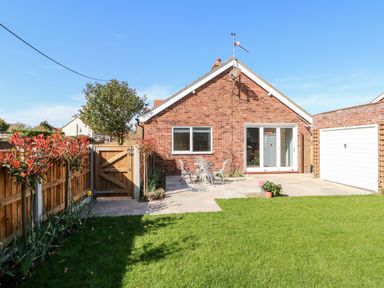 A backyard with lawn a patio with metal chairs and table a wooden fence a brick house with a garage at Sunnyside in Wells-Next-The-Sea