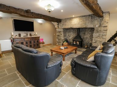 A living room with two black leather sofas a wooden coffee table a stone fireplace with a stove and a wooden cabinet with a television above at Hen Dy Craig Yr Ronwy Capel Celyn near Bala