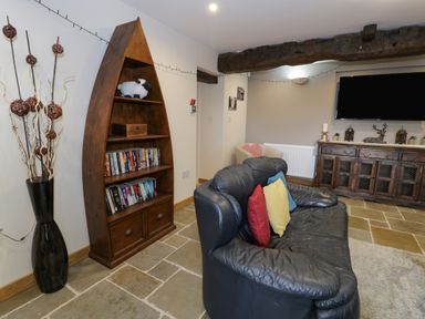 A living room with a black leather sofa with colorful cushions a wooden bookshelf shaped like a boat a vase with decorative sticks and a wooden cabinet with decorations and a TV on the wall at Hen Dy Craig Yr Ronwy in Capel Celyn near Bala