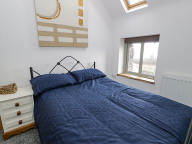A bedroom with a bed covered in blue bedding a nightstand with drawers and a woven basket a window with a blind and a skylight at Hen Dy Craig Yr Ronwy in Capel Celyn near Bala