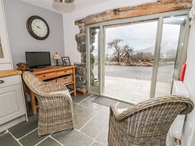 A room with two wicker chairs a wooden table with a small television lamp and photo frame stone floor and large glass doors opening outside at Hen Dy Craig Yr Ronwy in Capel Celyn near Bala