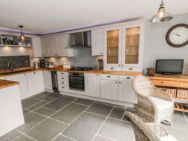 A kitchen with white cabinets wooden countertops a gas stove and oven a wicker chair and a wall clock at Hen Dy Craig Yr Ronwy in Capel Celyn near Bala