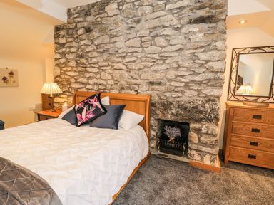 A bedroom with a wooden bed and white bedding against a stone wall with a small fireplace next to a wooden dresser with a mirror at Hen Dy Craig Yr Ronwy in Capel Celyn near Bala
