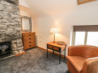 A room with a stone fireplace, wooden chest of drawers with a mirror, side table with a lamp, and a brown armchair at Hen Dy Craig Yr Ronwy in Capel Celyn near Bala