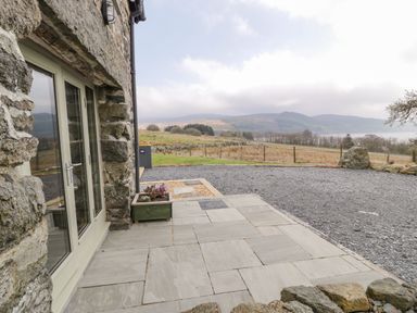 A stone-paved patio outside a stone building with glass doors and a planter with flowers in a rural area with hills in the background at Hen Dy Craig Yr Ronwy in Capel Celyn near Bala