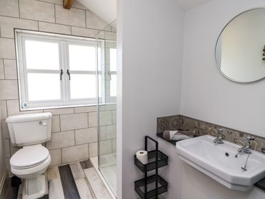 A bathroom with a white toilet near a window and a shower enclosure with a glass door a white sink with two taps a round mirror and a black metal stand holding a roll of toilet paper at Holida Hoose in Belford