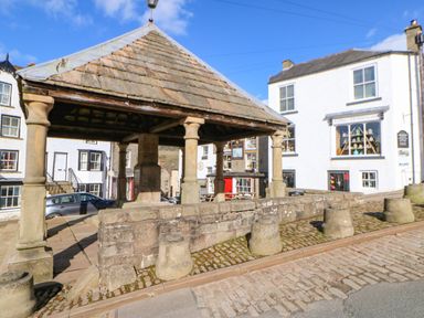A stone pavilion with pillars on a cobblestone street near white buildings with storefronts at 1 The Stables in Alston