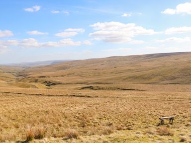 A bench in a wide open grassy landscape with hills and a partly cloudy sky at 1 The Stables in Alston
