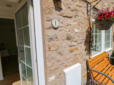 A stone exterior wall with a wooden bench and hanging flower basket at 1 The Stables in Alston