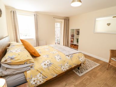 A bedroom with a bed with floral yellow bedding a bookshelf with books and wood and a wicker chair at 1 The Stables in Alston