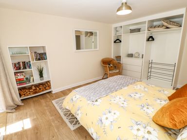A bedroom with a bed with yellow floral bedding a white bookshelf with books and firewood a wicker chair a white wardrobe and a mirror on the wall at 1 The Stables in Alston