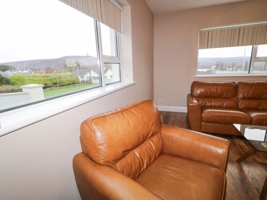 A living room with brown leather armchairs and sofas near windows overlooking a residential area at Loughanure Lake View in Loughanure County Donegal