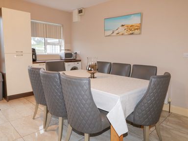 A dining area with a rectangular table covered with a tablecloth surrounded by eight gray quilted chairs a glass vase is on the table a kitchen area with appliances including a microwave and washing machine is in the background a framed beach landscape picture is on the wall at Loughanure Lake View in Loughanure County Donegal