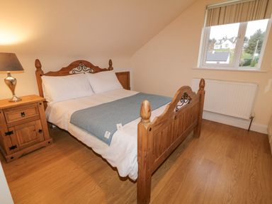 A bedroom with a wooden bed with white bedding and a blue blanket a wooden nightstand with a lamp and a window showing houses outside at Loughanure Lake View in Loughanure County Donegal