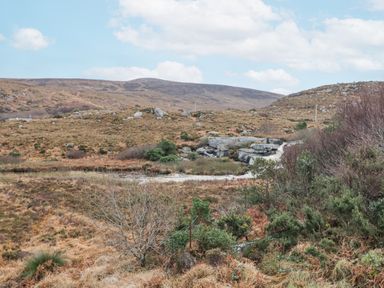 A rocky hillside with brown grass and sparse bushes under a partly cloudy sky at Loughanure Lake View in Loughanure County Donegal