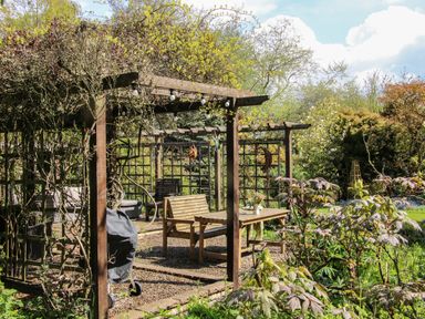 A garden seating area with wooden benches and table under pergolas surrounded by plants and trees at The Garden Lodge in Upton Cressett near Bridgnorth