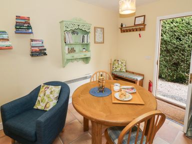 A dining area with a round wooden table set with food and a French press a blue armchair with a cushion a wooden bench with a cushion bookshelves on the wall and a door leading outside at The Garden Lodge in Upton Cressett near Bridgnorth