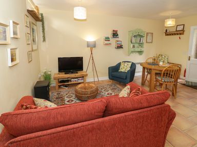 A living room with a red sofa a blue armchair a wooden dining table with chairs a television on a wooden stand and wall-mounted shelves at The Garden Lodge in Upton Cressett near Bridgnorth