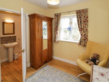 A room with a wooden wardrobe a window with floral curtains a yellow armchair with a teddy bear and a view into a bathroom with a sink and mirror at The Garden Lodge in Upton Cressett near Bridgnorth