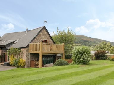 A stone house with a wooden balcony and green lawn with trees and hills in the background at Pentre Barn in Llantilio Pertholey near Mardy