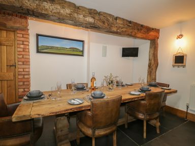 A dining room with a wooden table set with dishes and glasses six leather chairs a wooden door exposed brick and a wooden beam on the ceiling a landscape painting and a small wall-mounted television at Pentre Barn in Llantilio Pertholey near Mardy