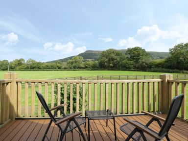 A wooden deck with two chairs and a small table overlooking a fenced grassy field with trees and hills in the background at Pentre Barn in Llantilio Pertholey near Mardy