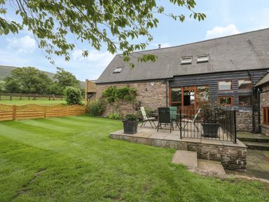 A garden with lawn chairs and table on a patio outside a stone and wood house at Pentre Barn in Llantilio Pertholey near Mardy