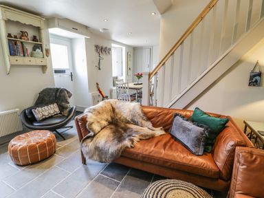 A living room with a brown leather sofa with pillows and a fur throw a black chair with a blanket and cushion a brown ottoman and a dining table with chairs near a staircase at Swn yr Afon in Beddgelert