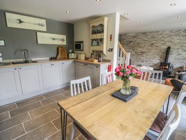 A kitchen and dining area with a wooden table and flowers in a vase and a fireplace with fire at Swn yr Afon in Beddgelert
