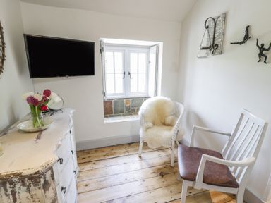 A small room with a mounted TV a distressed white dresser with flowers two chairs and a window at Swn yr Afon in Beddgelert