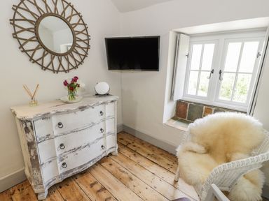 A corner of a room with a distressed white wooden dresser a round wall mirror a wall-mounted TV a window with shutters and a white wicker chair with a fur throw at Swn yr Afon in Beddgelert