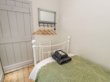 A small bedroom with a white metal bed frame a green blanket folded towels on the bed wooden floor a gray door a wall mirror with hooks and wooden hangers at Swn yr Afon in Beddgelert