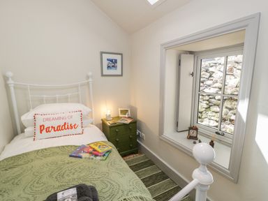 A small bedroom with a white metal bed a green blanket and a cushion with text a bedside table with a lamp and a window showing a stone wall at Swn yr Afon in Beddgelert