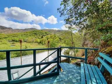 An outdoor wooden deck with a bench and a railing overlooking a hillside covered with trees at Swn yr Afon in Beddgelert