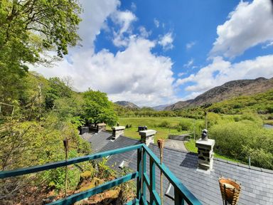 A balcony overlooking a roof with chimneys and a green valley with hills under a blue sky at Swn yr Afon in Beddgelert