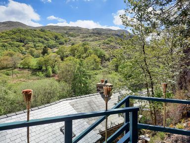 View of a hillside with trees and a slate roof seen from a blue balcony with tiki torches at Swn yr Afon in Beddgelert