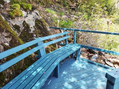 A blue wooden bench on a deck next to a mossy rock wall with greenery surrounding at Swn yr Afon in Beddgelert