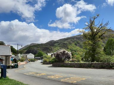 A street with buildings and a bus stop area near hills under a partly cloudy sky at Swn yr Afon in Beddgelert