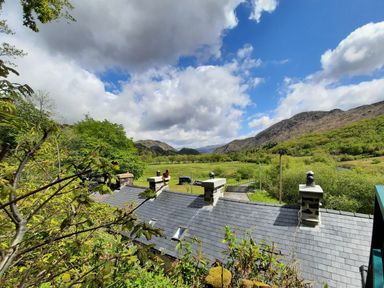 A view of a slate roof with chimneys trees fields and mountains under a cloudy sky at Swn yr Afon in Beddgelert
