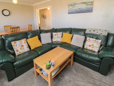 A living room with a green leather corner sofa with cushions a wooden coffee table with plants and magazines and a dining table with chairs at Island View Cottage in High Hauxley near Amble
