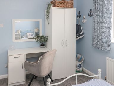 A bedroom corner with a white desk and mirror a grey chair a white wardrobe and blue patterned curtains at Island View Cottage in High Hauxley near Amble