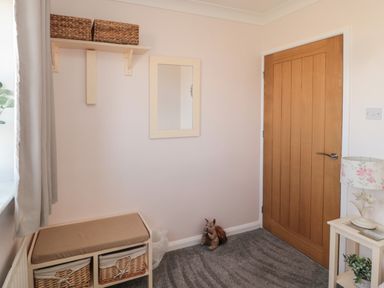 A small room with a wooden door a bench with wicker baskets a mirror on the wall a toy squirrel a lamp on a small table and a window with curtains at Island View Cottage in High Hauxley near Amble