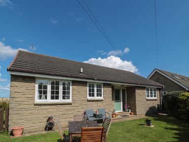A stone bungalow with white windows and a green door a wooden table and chairs on grass outside Island View Cottage High Hauxley near Amble