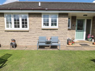 The exterior of a stone house with two windows a green door a blue bench with a small table and several potted plants on a stone patio with a grass lawn