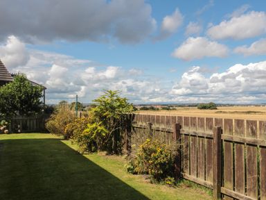 A backyard with grass and shrubs next to a wooden fence and open fields beyond at Island View Cottage in High Hauxley near Amble