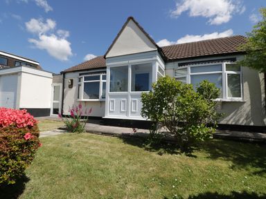 A single story house with a bay window and porch surrounded by a grassy garden with bushes and flowers at Garden View in Quintrell Downs near Newquay