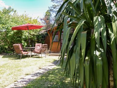 A garden with a large plant in the foreground a path leading to a seating area with four chairs and a red umbrella and a wooden shed in the background at Garden View in Quintrell Downs near Newquay